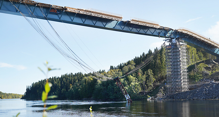 Bilden är tagen utomhus. Vi ser en bro som håller på att byggas över vatten. En stor bit av metall hänger ner från bron i vajrar. Den hänger nere vid vattnet.