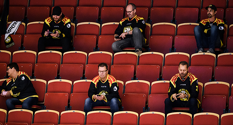 Några män sitter på läktaren. De kollar på en match i ishockey. De har stort avstånd mellan varandra.