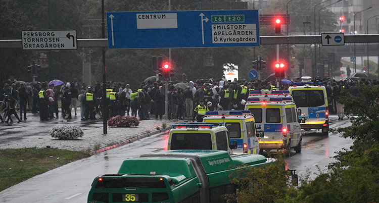 Flera polisbilar och poliser står på en regnig gata. Bakom dem syns en massa människor på långt håll.