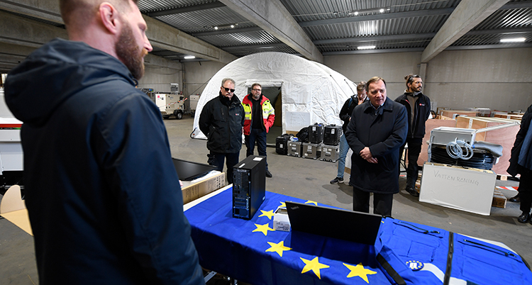 Flera personer står inne i en stor lokal. I mitten står statsministern Stefan Löfven. En EU-flagga ligger på ett bord.