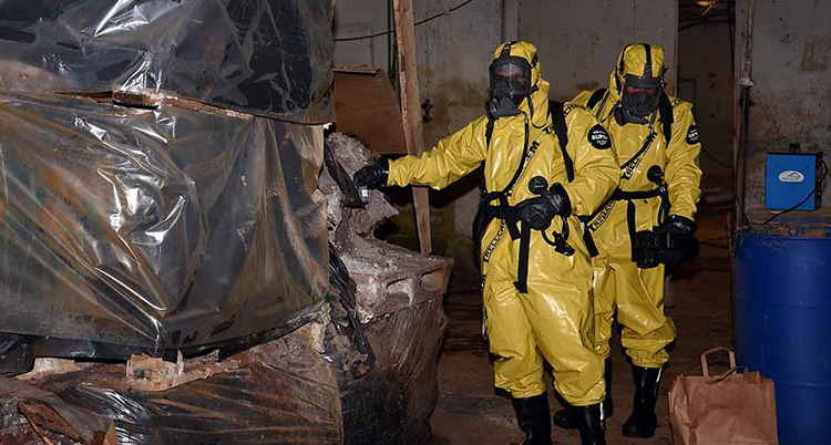 Två personer i gula skyddskläder och gasmasker står vid stora inplastade föremål i en källarlokal.