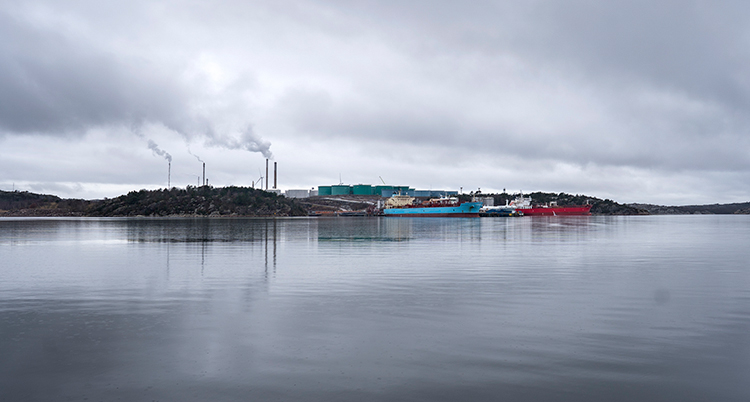 Flera stora båtar syns på havet. Fabriken syns i bakgrunden. Det ryker från fabrikens skorstenar.