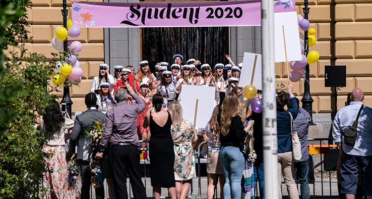 Ett gäng med studenter firar på en trappa med sina mössor på. Folk står runt dem med ballonger och skyltar.