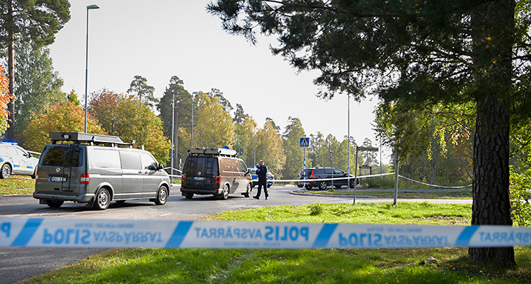 Bilden är tagen utomhus. Poliserna har satt upp ett band för att spärra av. Bakom bandet syns flera polisbilar. Runt vägen finns gräsmattor och träd.