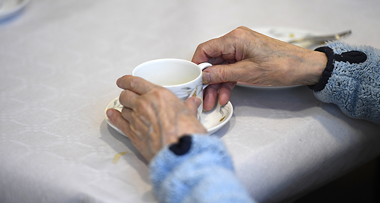 Vi ser händerna på en gammal människa. Händerna håller om en kaffekopp.