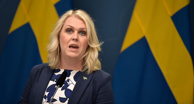 Lena Hallengren under en presskonferens. Hon står framför två svenska flaggor. Det ser ut som att hon håller på att säga något.