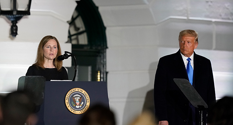 Amy Coney Barrett står på en scen och pratar. Hon står vid en talarstol och pratar i en mikrofon. Till höger i bilden står Donald Trump.