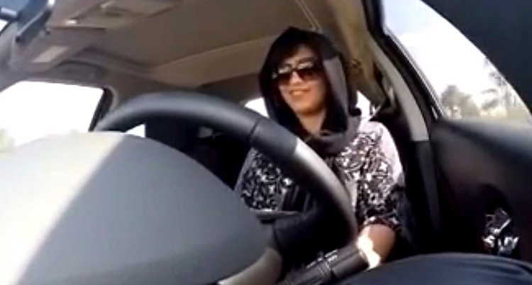 Vi ser en kvinna som kör bil. Hon har solglasögon och en svart slöja över håret.