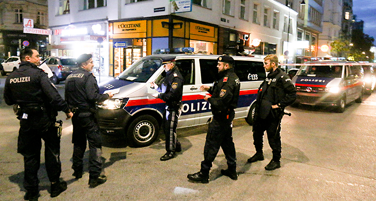 Fem poliser står på en gata mitt i en stad. Poliserna har svarta kläder. Flera polisbilar finns också på gatan.