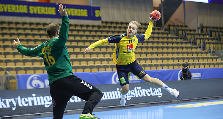 Från en match i handboll. En spelare i Sveriges lag hoppar högt i luften. Han har bollen i handen och ska skjuta. Målvakten gör sig beredd att rädda bollen.