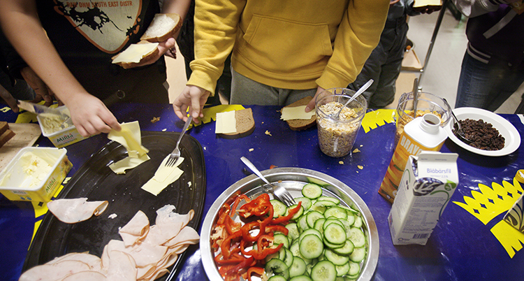 bild tagen ovanifrån ett bord med mat, gurka, skinka, paprika. Vi ser också armar från flera barn som lägger pålägg på mackor. Bilden är från barn som äter frukost på en skola.