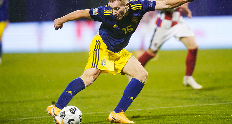 En fotbollsspelare i svensk landslagsdräkt i blått och gult dribblar med en fotboll.