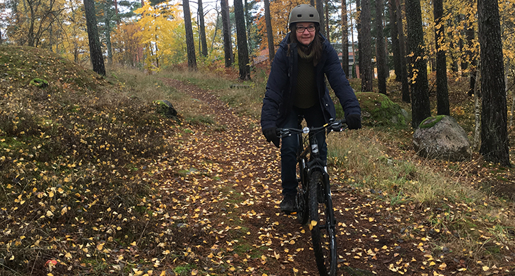 En kvinna i mörka kläder och cykelhjälm cyklar i en höstfärgad skog på en cykel