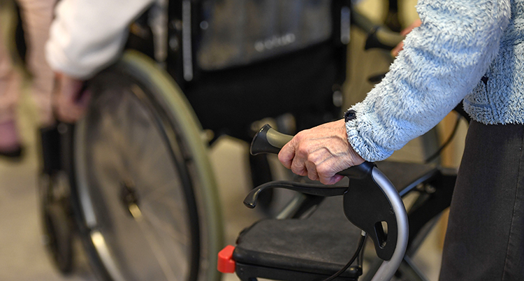 Ett par händer håller i ett handtag på en rollator. Två hjul på en rullstol syns också.