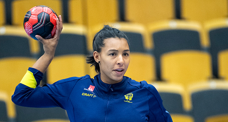 En kvinna med mörkt hår i tofs, blå träningströja och koncentrerad blick, håller en handboll som hon är beredd att kasta med höger hand bakifrån huvudet