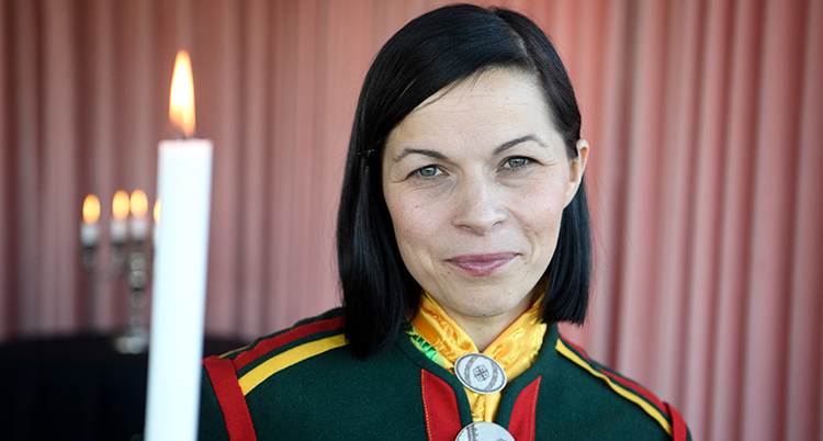 Närbild på Labba. Hon är klädd i en samiska dräkt i gult, grönt. rött och gult. Bredvid henne får ett levande ljus.