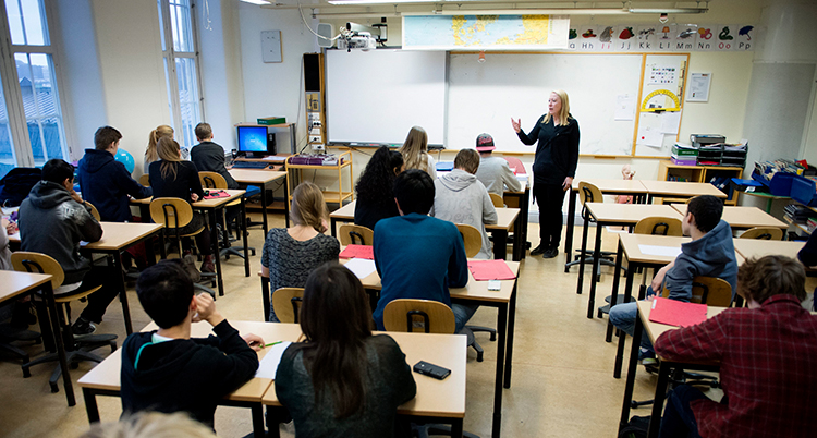 Läraren står längst fram vid tavlan i ett klassrum.