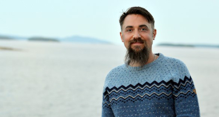 En man med mörkt hår och skägg och en mönsterstickad tröja står på en strand