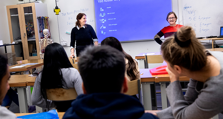 Ett klassrum i en skola. Två lärare står framför en blå datorskärm. Man ser ryggen på några elever som tittar på dem.