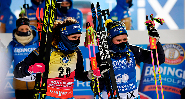 Hanna och Elvira står bredvid varandra. De håller i sina skidor och stavar. De är glada. De har munskydd på sig.