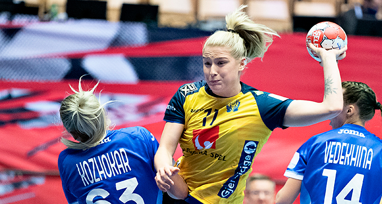 Från en match i handboll. En svensk spelare kastar sig fram för att skjuta. Hon har bollen i handen. En motståndare försöker stoppa henne.