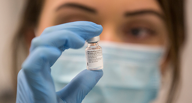 En kvinna håller i en liten flaska med vaccin i. Hon har blå plasthandskar på sig och ett ljusblått skydd för munnen.