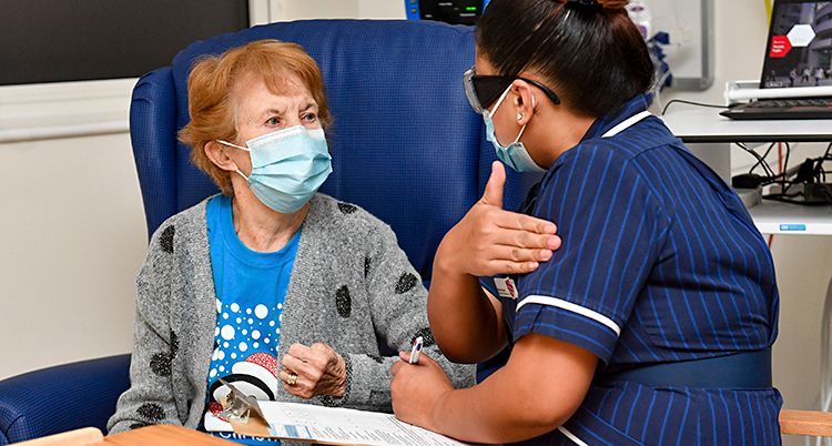 Bilden är tagen på ett sjukhus. Två personer är på bilden. Till vänster sitter en 90-årig kvinna. Till höger sitter en sjuksköterska.