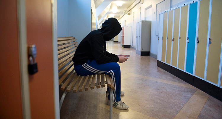 en ensam elev med huva uppdragen över huvudet sitter i en korridor i en skola och tittar i sin mobil