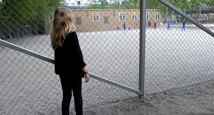 en flicka med svarta kläder och långt hår står vid ett gallerstaket och tittar in mot en skolgård där barn leker