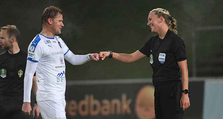 En spelare i vit tröja och Tess Olofsson i svarta domarkläder. Båda ler och stöter nävarna mot varandra för att tacka efter matchen.