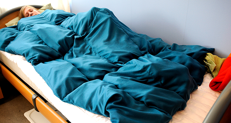 En person ligger i en säng under ett turkost täcke, det är ett tyngdtäcke