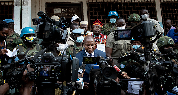 Bilden är tagen utomhus i landet Centralafrikanska republiken. Landets president Faustin Archange Touadéra blir intervjuad. Han står framför många kameror och mikrofoner. Bakom honom står många personer, bland annat soldater från FN.