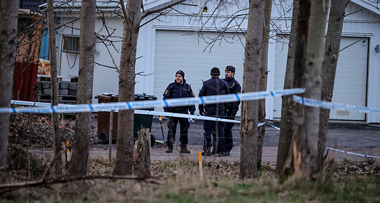 Tre poliser står utomhus. De står vid ett vitt garage. Poliserna har satt upp vita och blå band mellan träd.