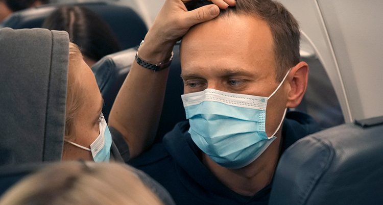 Navalnyjs fru håller en hand på hans panna. de tittar på varandra. De sitter i varsin flygplansstol bredvid varandra.