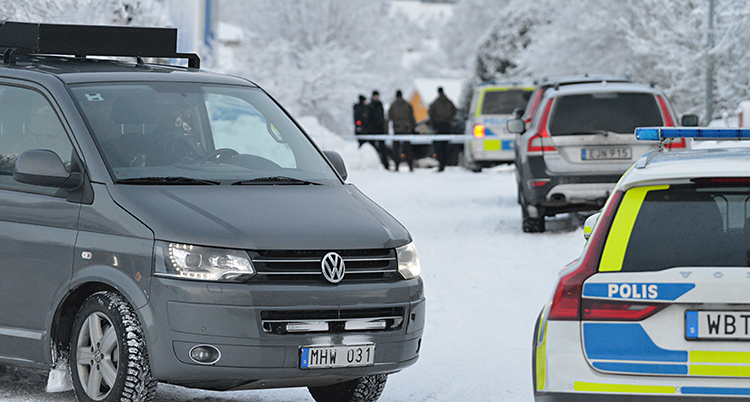 Vi ser en gata. Det är snö på gatan. Flera polisbilar står på gatan. Och längre bort står några poliser.