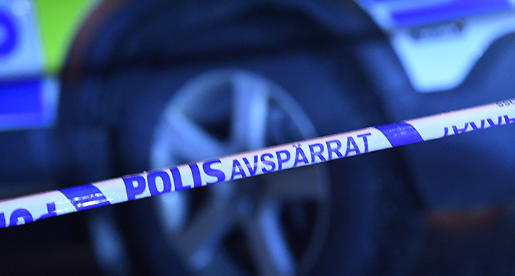 Vi ser ett blått och vitt band. Där står det Polis avspärrat. I bakgrunden syns en polisbil.