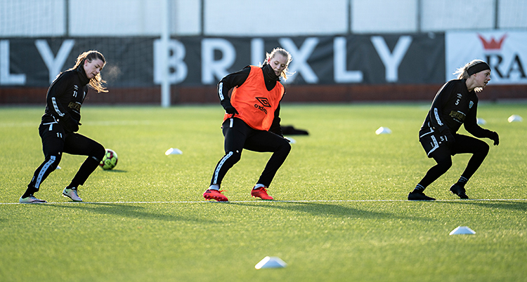 Tre kvinnor är på en fotbollsplan. De tränar. De gör en övning där de springer.