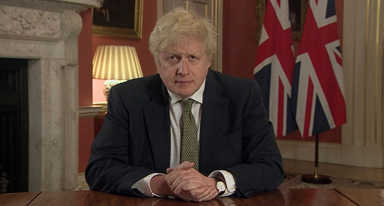 Han sitter vid ett skrivbord. Bakom honom syns Storbritanniens flagga.