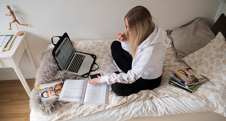 En ung flicka sitter i en säng. Framför henne står en dator. På sängen liggen en uppslagen bok.
