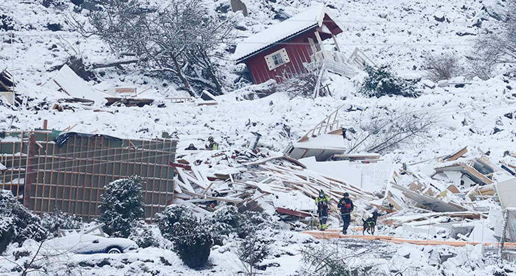 Ett rött hus syns i den snöiga raskanten. Räddningsarbetare går i det snöiga rasområdet.