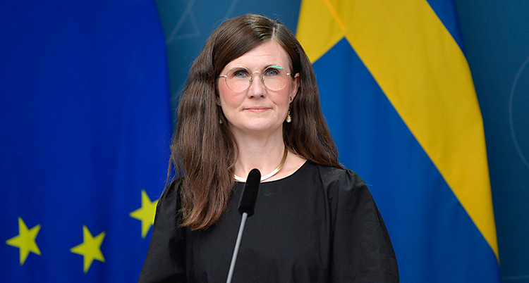 Hon har långt mörkt hår och glasögon. Hon står vid en mikrofon. I bakgrunden är det Sveriges flagga och EUs flagga.