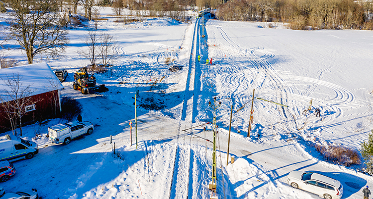 En bild som är tagen uppifrån luften. Nedanför ser vi en väg för bilar. Ett spår för tåg går rakt över vägen. Ett hus och några bilar syns också. Och ett fält med snö och träd. Det är ute på landet.