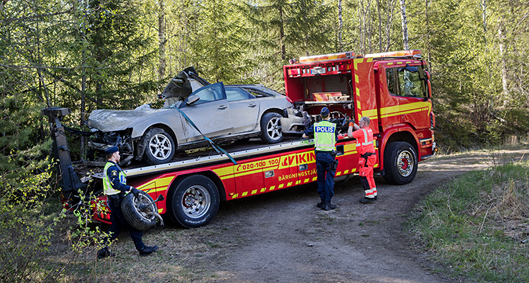 Vi ser en stor och röd bärgningsbil. Den har lastat upp en personbil som är grå. Personbilen är mycket skadad.