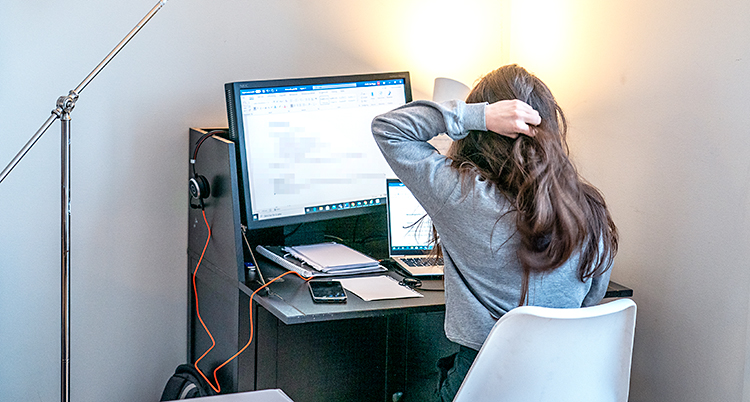 En flicka sitter hemma vid ett skrivbord. Hon har en dator framför sig. Vi ser henne bakifrån. Hon har långt hår.