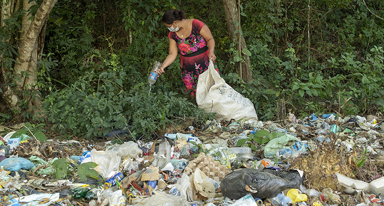 En kvinna står i ett skogsbryn. Hon har en stor påse i ena handen och håller en plastflaska i den andra. Framför henne ligger massor av sopor på marken.