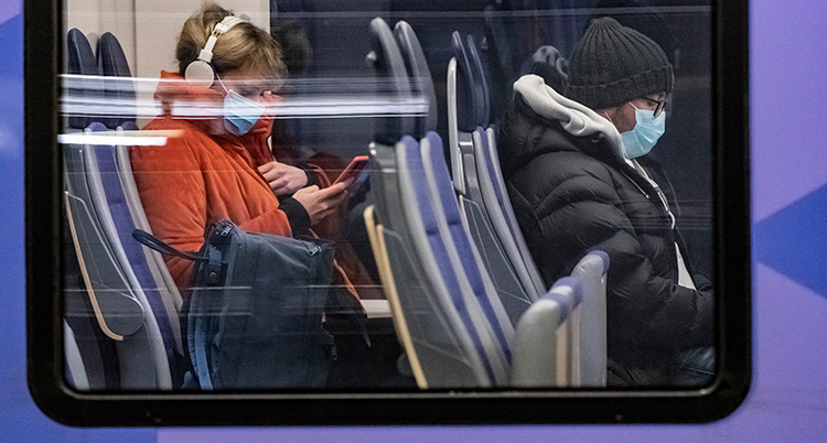 Två resenärer med munskydd syns genom ett fönster på en buss eller ett tåg.