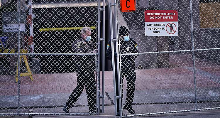Bilden visar en stor gallergrind utanför ett hus. Två poliser står innanför gallret. De har munskydd på sig.