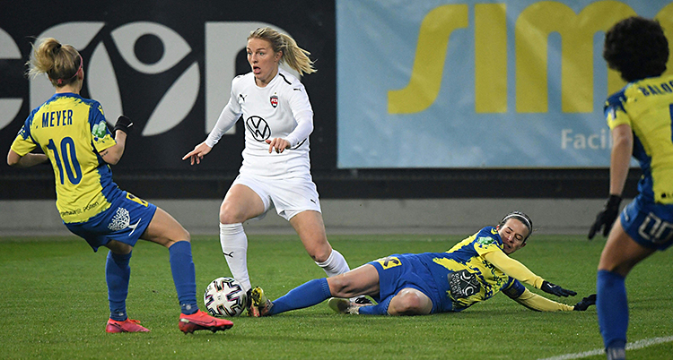 En match i fotboll. En kvinna med vita kläder har bollen. Det andra laget har blå och gula dräkter.