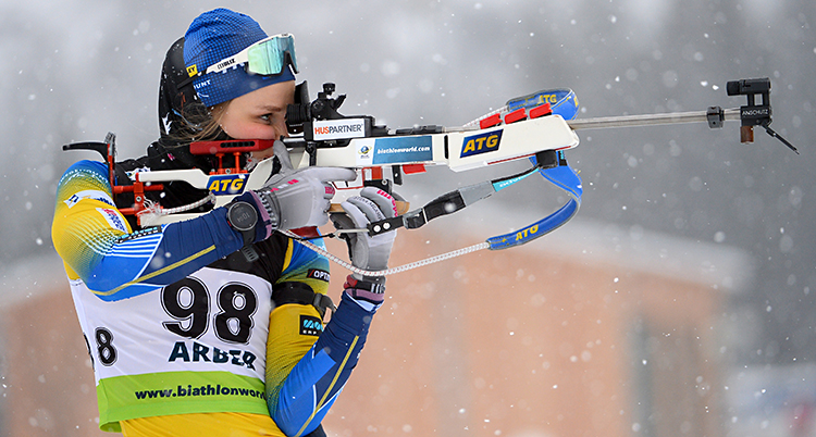 Stina Nilsson på en tävling. Hon siktar med sitt gevär. Hon har en gul och blå dräkt. Det snöar litegrann.