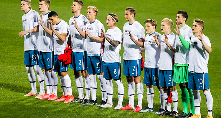 Norges lag står på en grön fotbollsplan. Spelarna står bredvid varandra. De har vita t-shirts med svart text på.